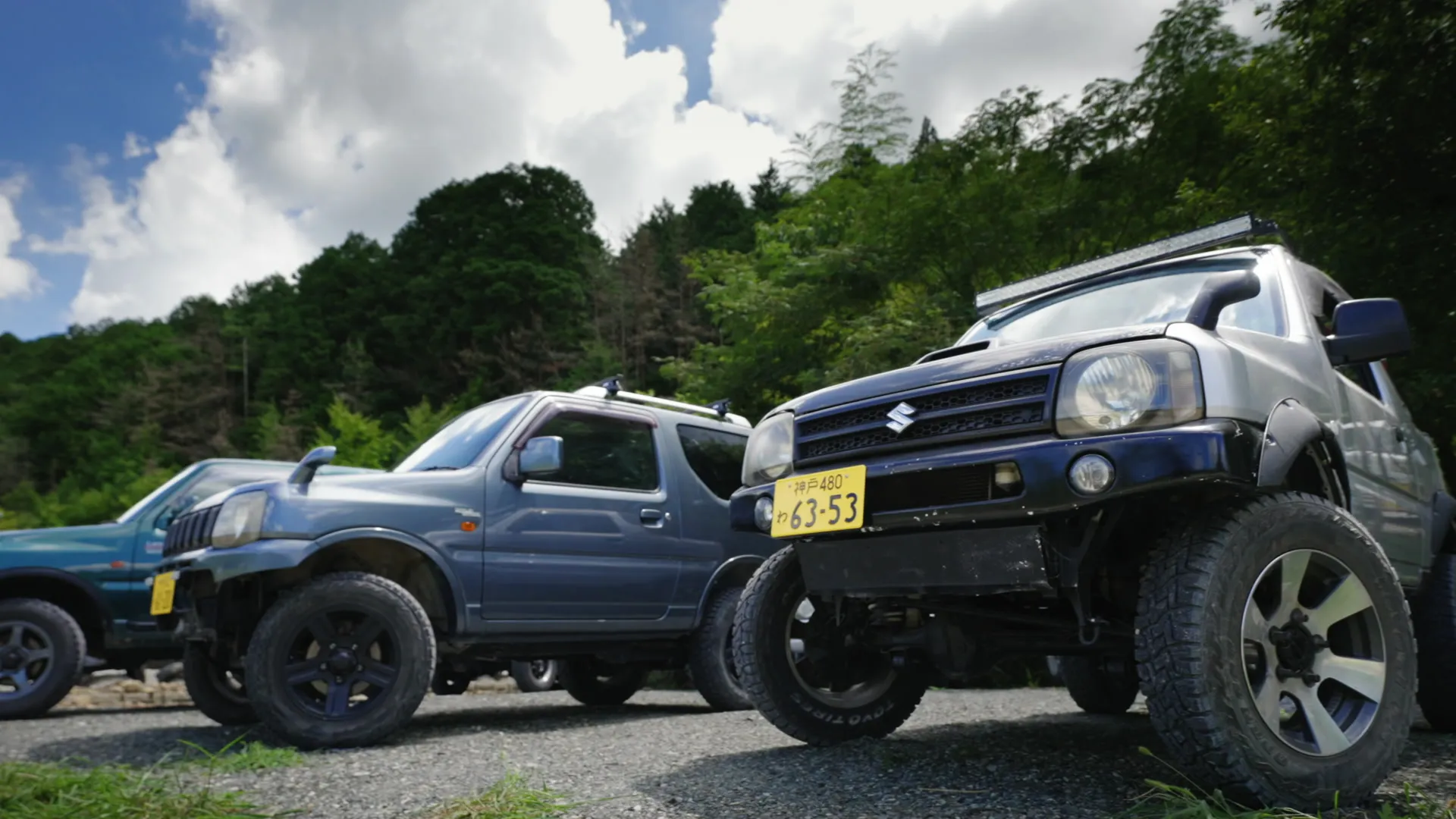 Three Jimnys lined up under a summer sky, the forest behind them
